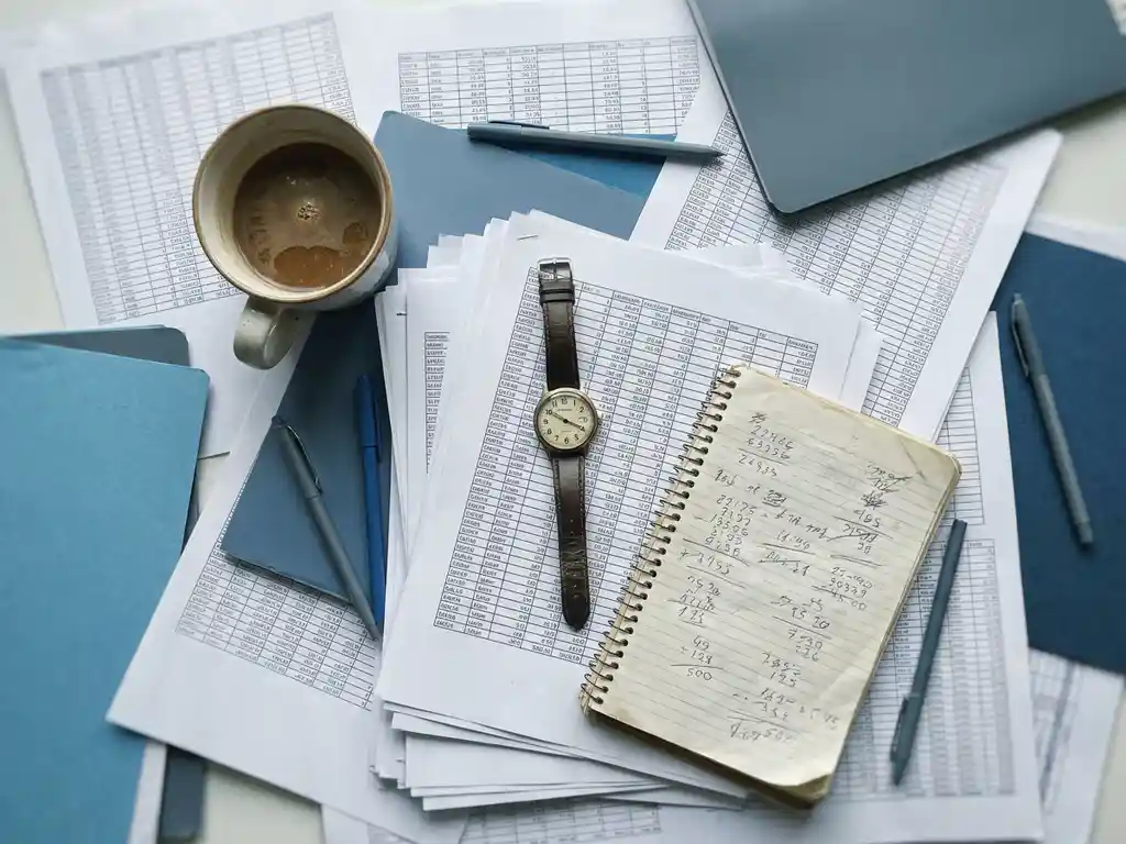 Overhead flat lay of a cluttered desk with stacked spreadsheets, handwritten notepad, forgotten coffee mug, and wristwatch symbolizing lost time.